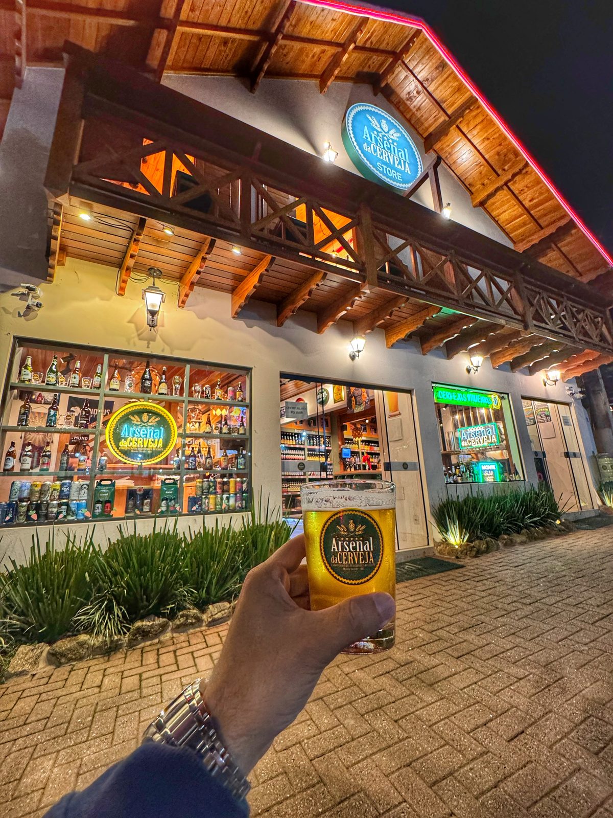 Fachada do Arsenal da Cerveja na Vila Germânica à noite, com letreiro iluminado e vitrines de cervejas