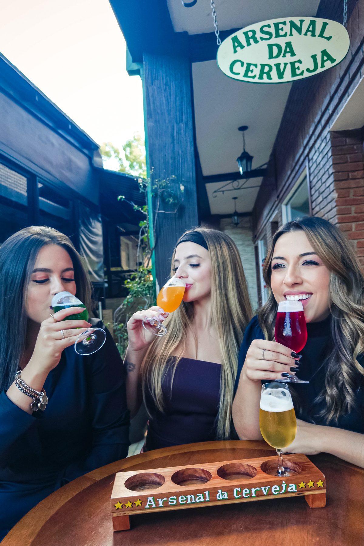 Três amigas brindando com as taças da régua de degustação na varanda do Arsenal da Cerveja
